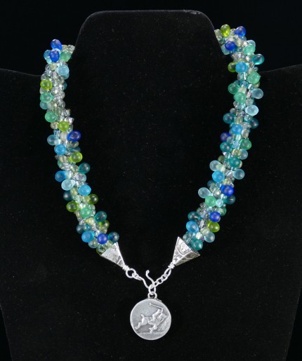 “Seafoam Polo Necklace” Overall length: 16", Polo pendant: 7/8" Diameter, Price: $550, Chisholm Ref. #: SBW-N10 | This one-of-a-kind silver & beadwork necklace is woven of glass beads: matte teardrops, Czech fire polished crystals, and seed bead, all in hues of the brilliant ocean. The silver end-caps are textured with wildflower leaves, and the focal piece features a polo player & pony both totally intent on the ball. The findings are all handcrafted in .960 silver.